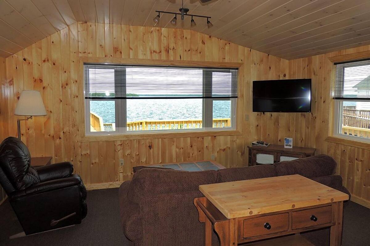 Interior acogedor de una cabaña con paredes de madera, ventilador de techo, televisor, sillón de cuero, sofá y una mesa de centro de madera.