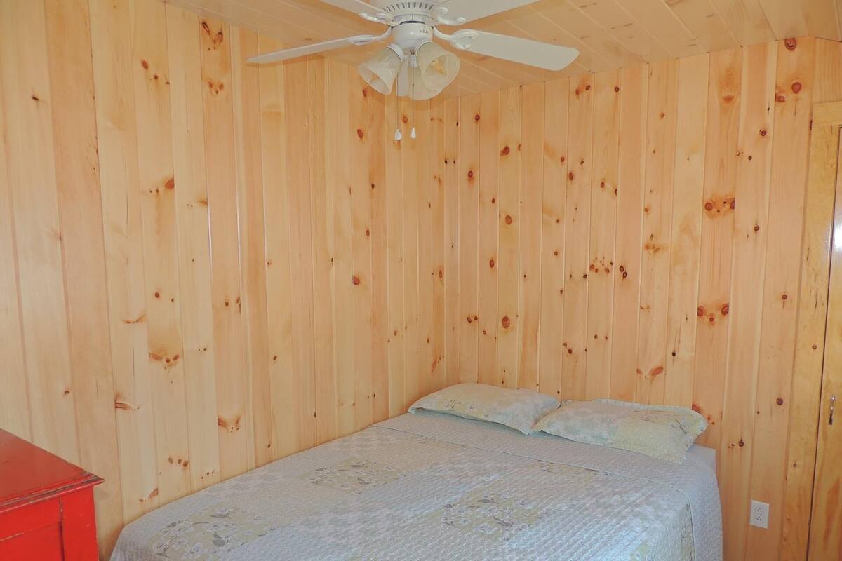 Una habitación con paredes de madera, una cama con ropa de cama floral, un ventilador de techo y un mueble rojo.