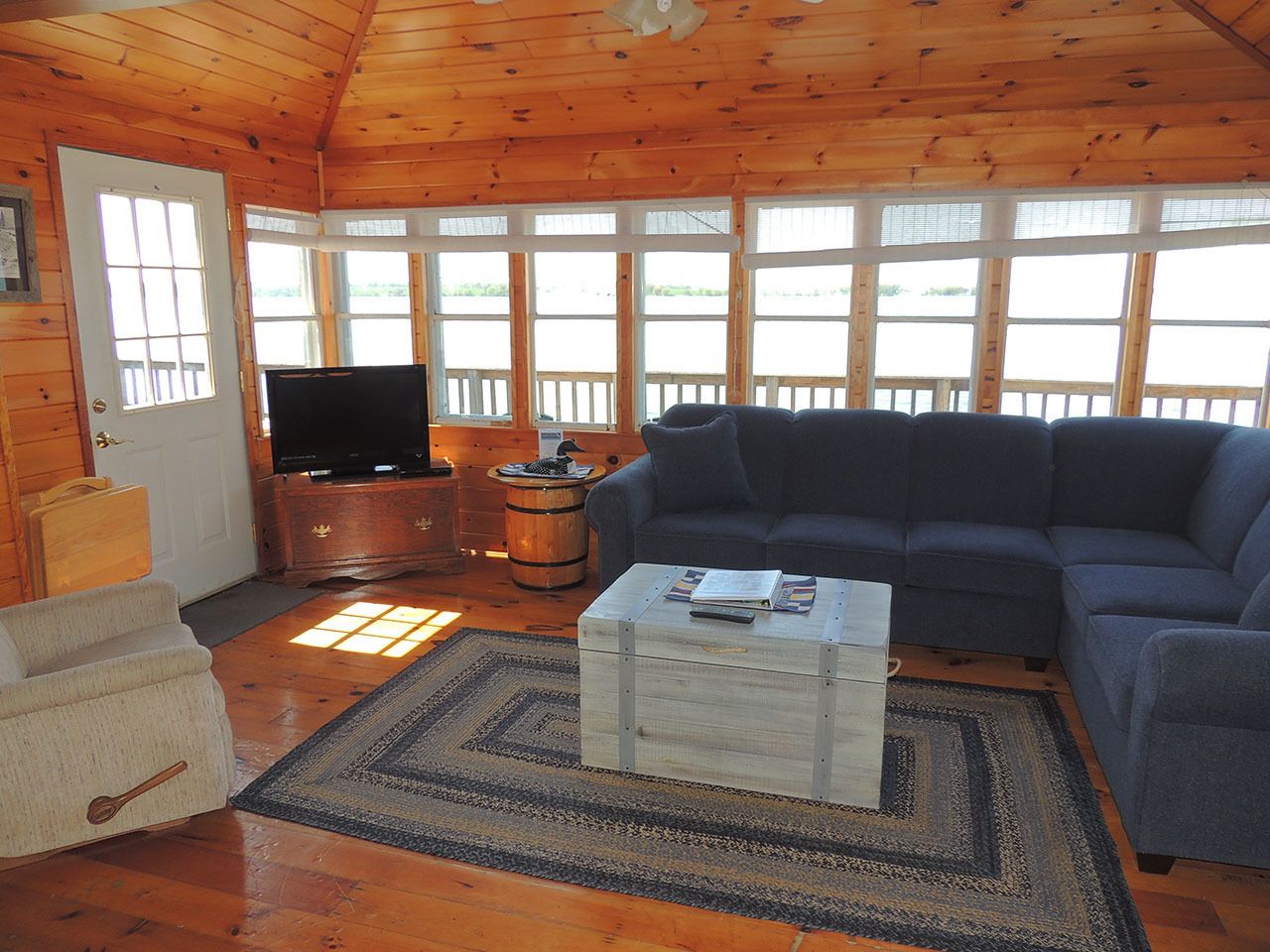 A cozy cabin interior with a wooden ceiling, a large window, a blue sofa, a television, and a wooden chest.