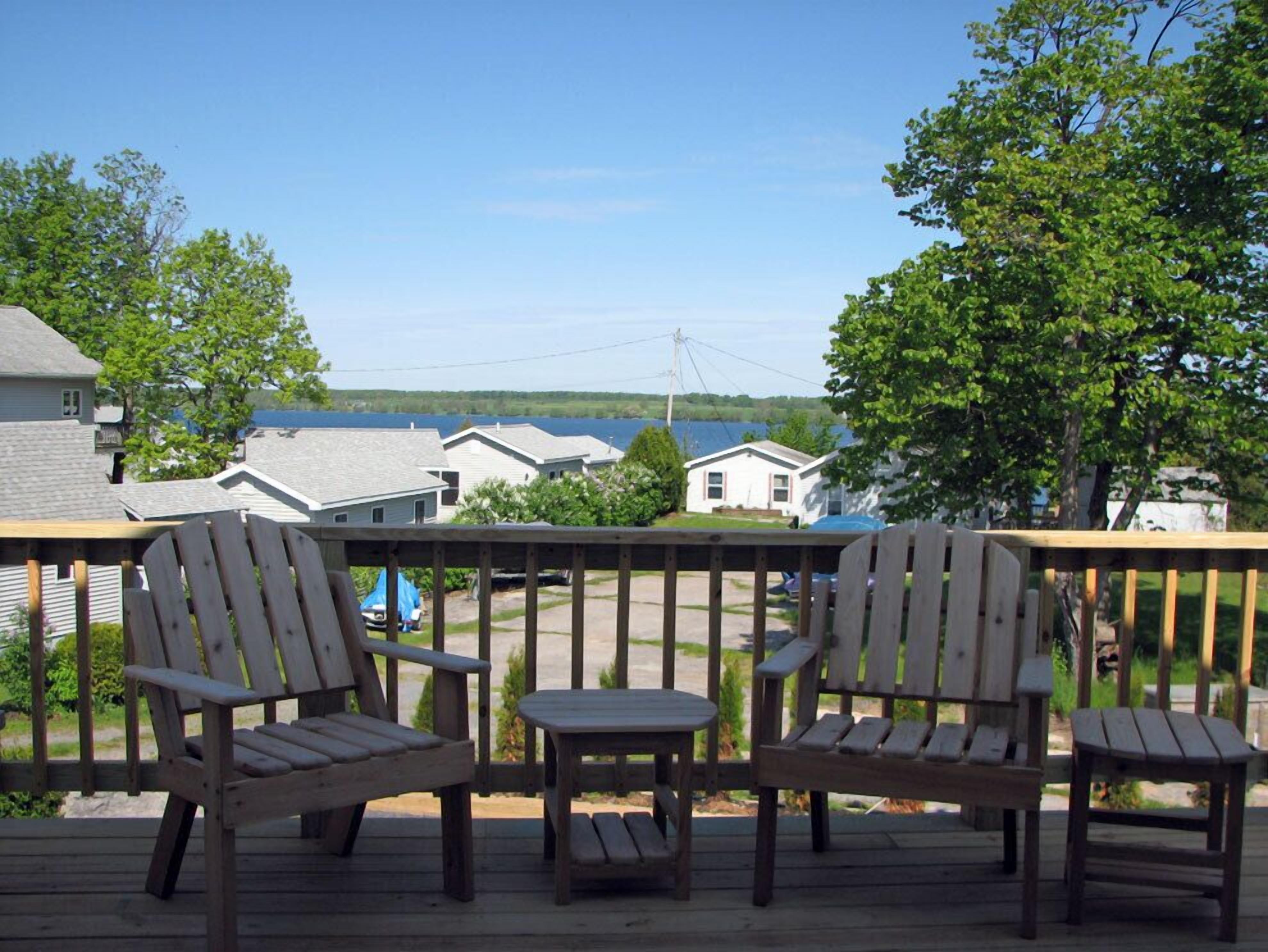 A wooden deck with two Adirondack chairs and a small table, overlooking a lake with houses in the background.
