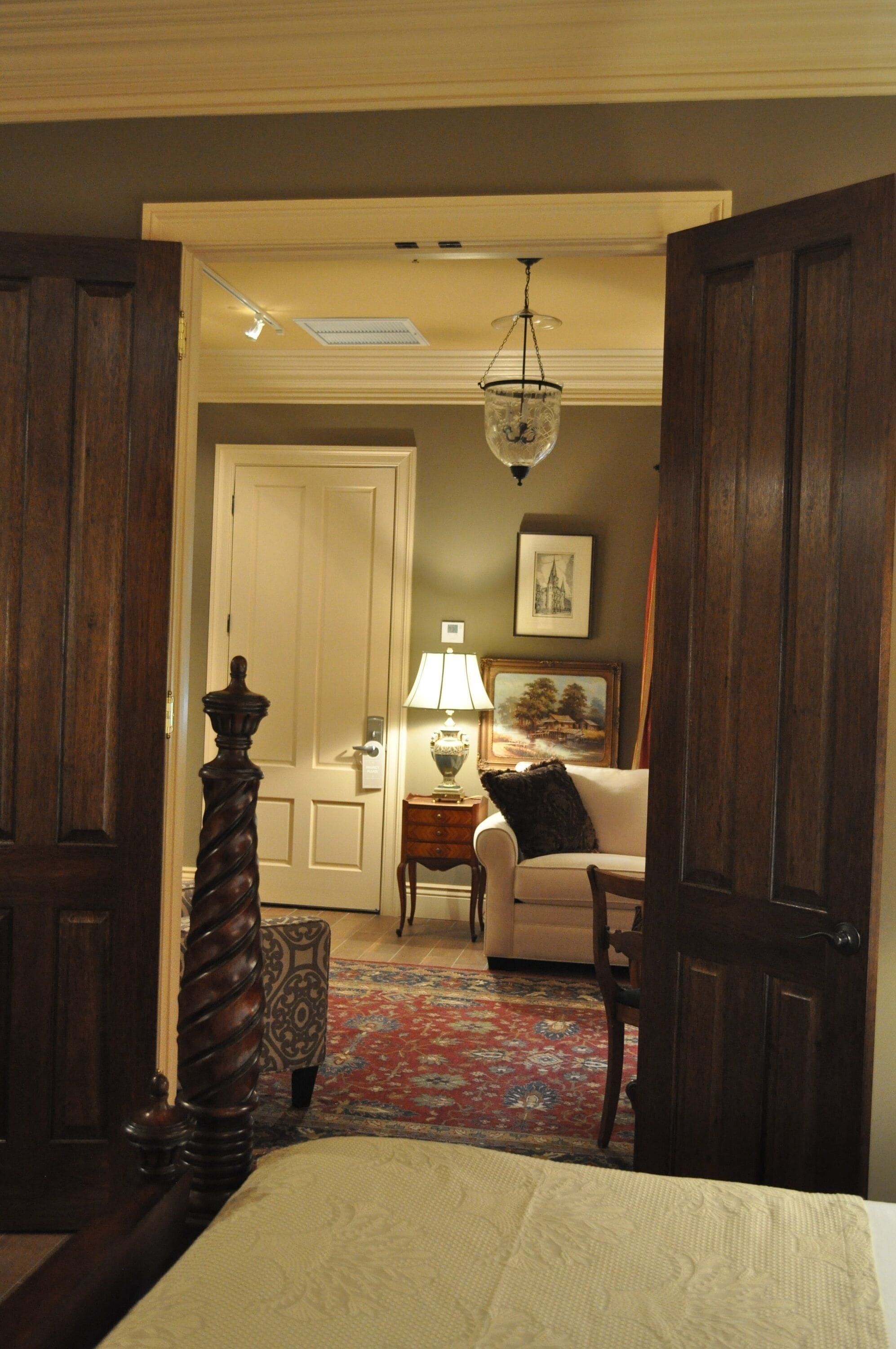 A hotel room with a bed, a wooden wardrobe, a nightstand with a lamp, a chair, and a painting on the wall.