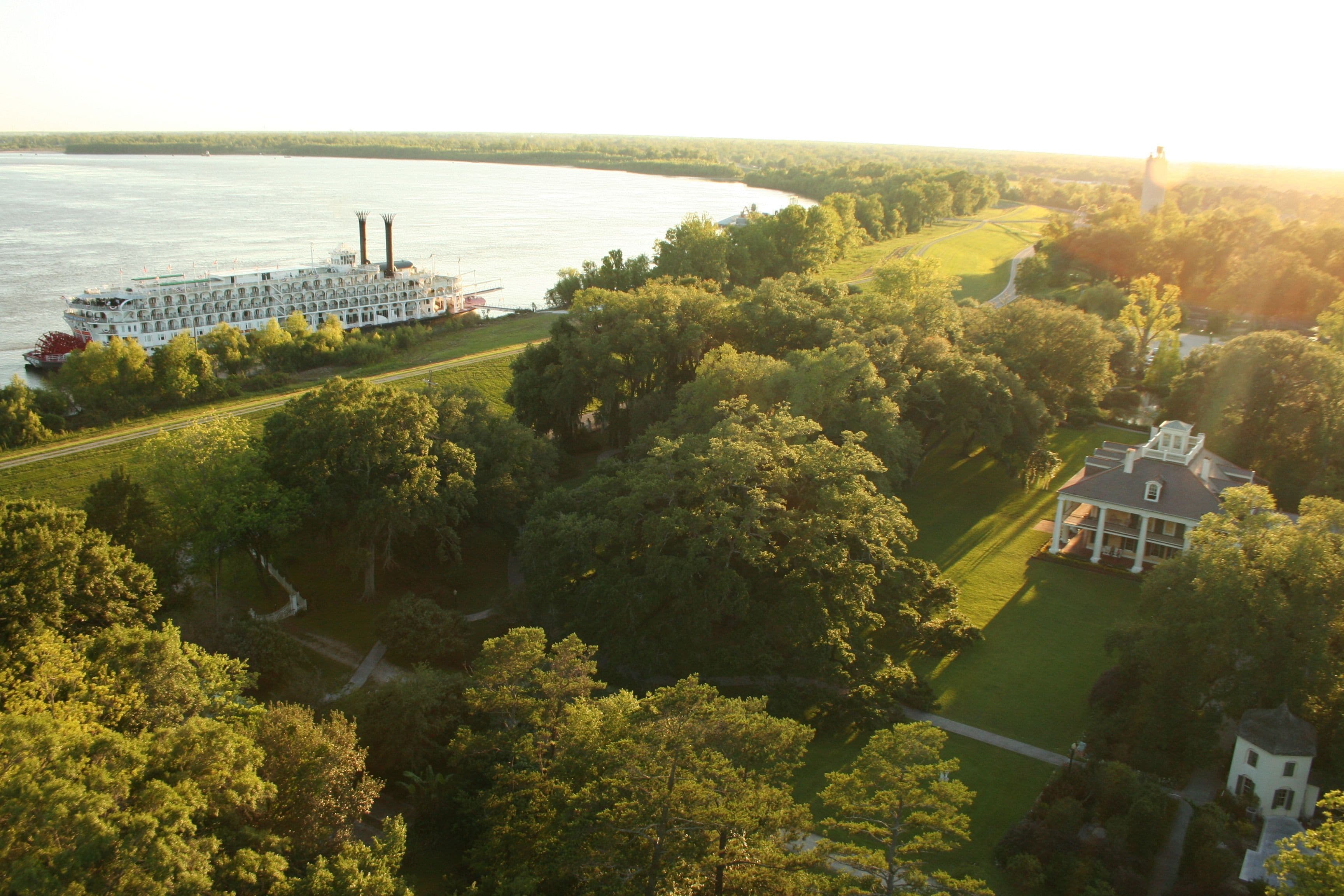 A riverboat docked near a river, surrounded by lush greenery and a large estate with a prominent house and smaller structures.