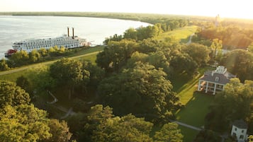 A riverboat docked near a river, surrounded by lush greenery and a large estate with a prominent house and smaller structures.