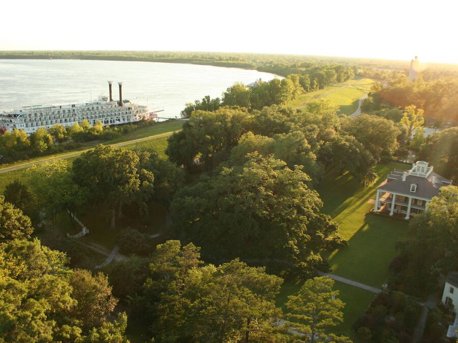 A riverboat docked near a river, surrounded by lush greenery and a large estate with a prominent house and smaller structures.