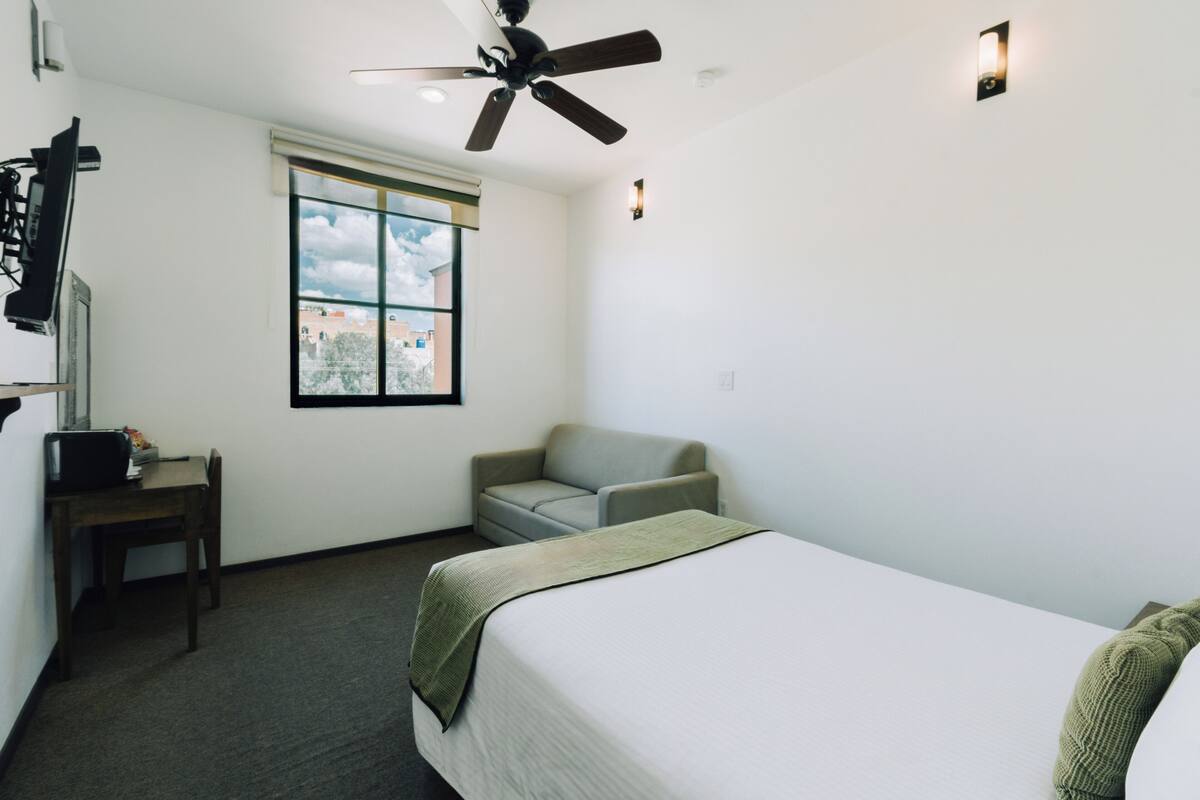 A hotel room with a bed, a desk, a chair, a ceiling fan, a window, and a view of the outside.