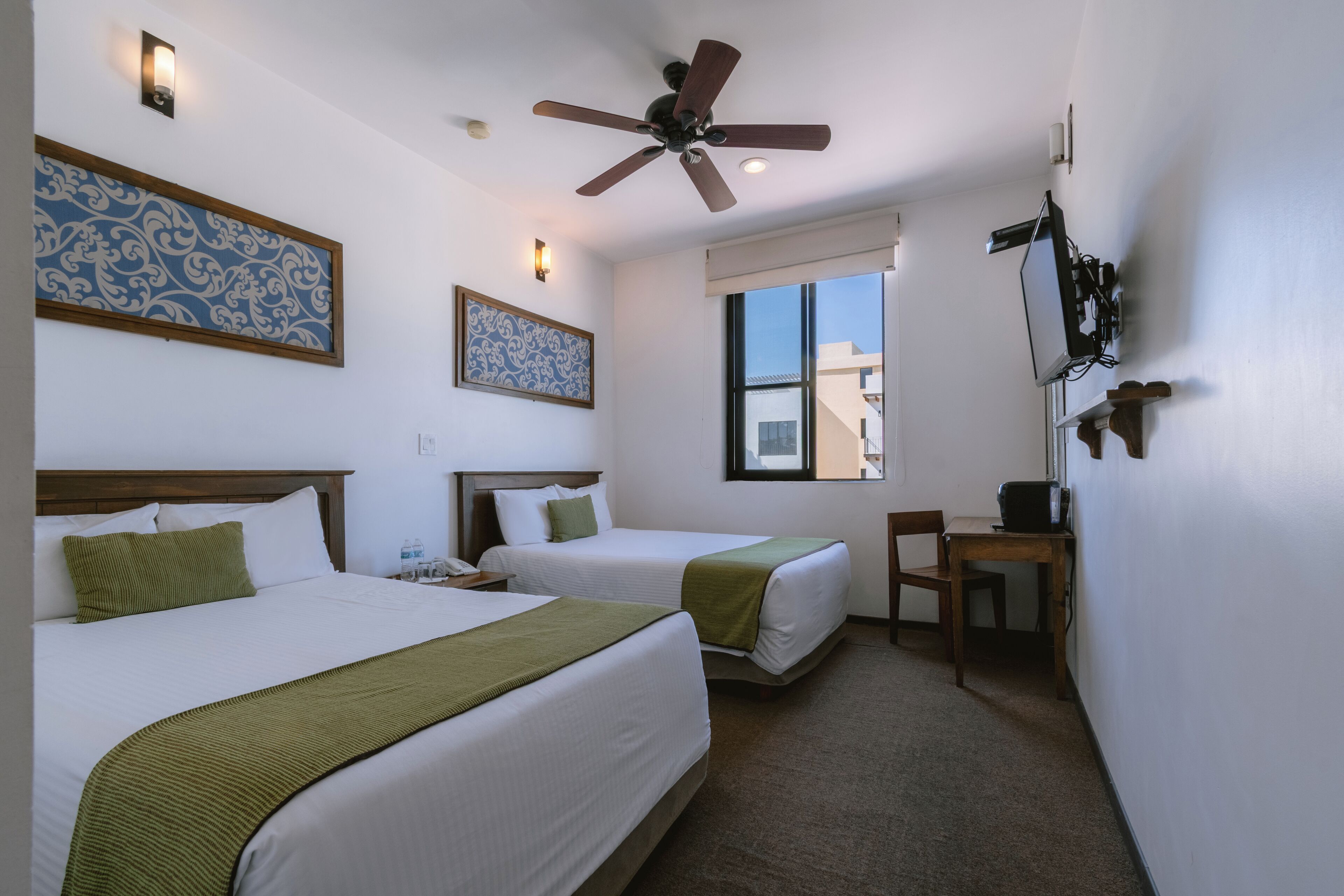 A hotel room with two beds, a ceiling fan, a desk, and a TV mounted on the wall.