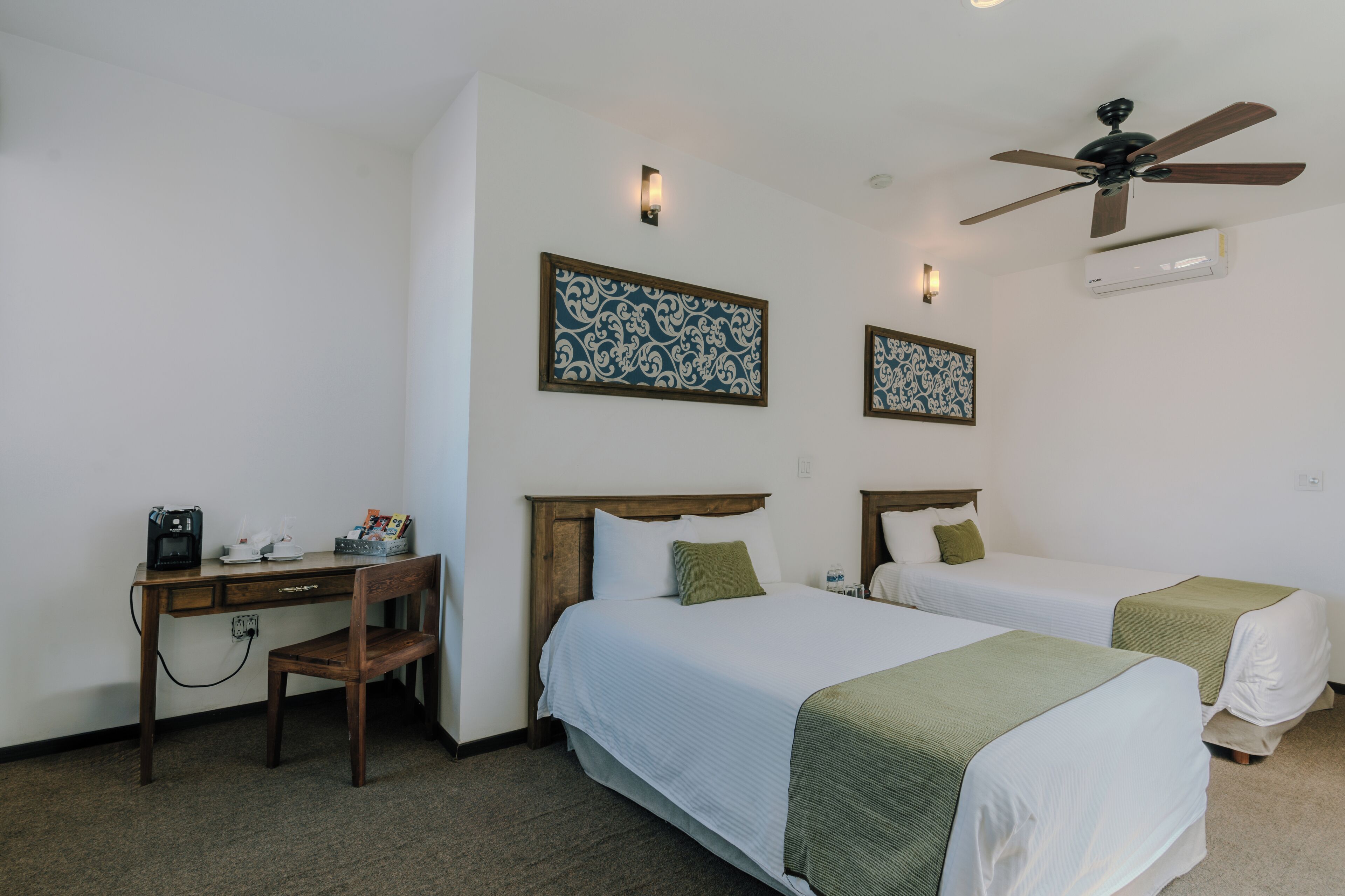 A hotel room with two beds, a desk, a chair, and a ceiling fan.