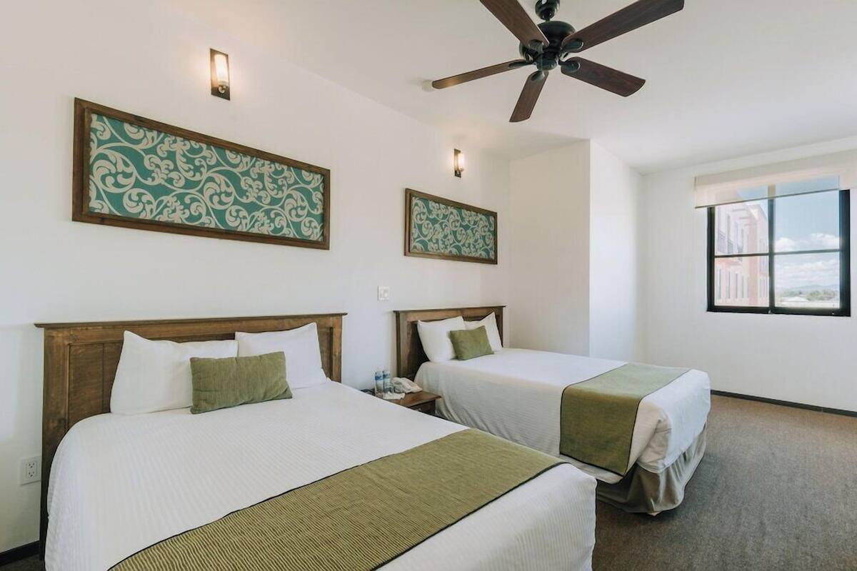 A hotel room with two beds, a ceiling fan, wall-mounted light fixtures, and a window with a view.