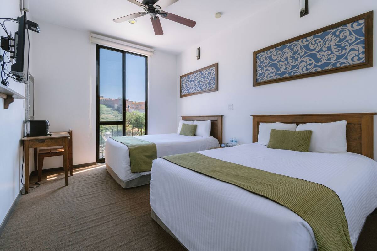 A hotel room with two beds, a desk, a ceiling fan, and a large window offering a view.