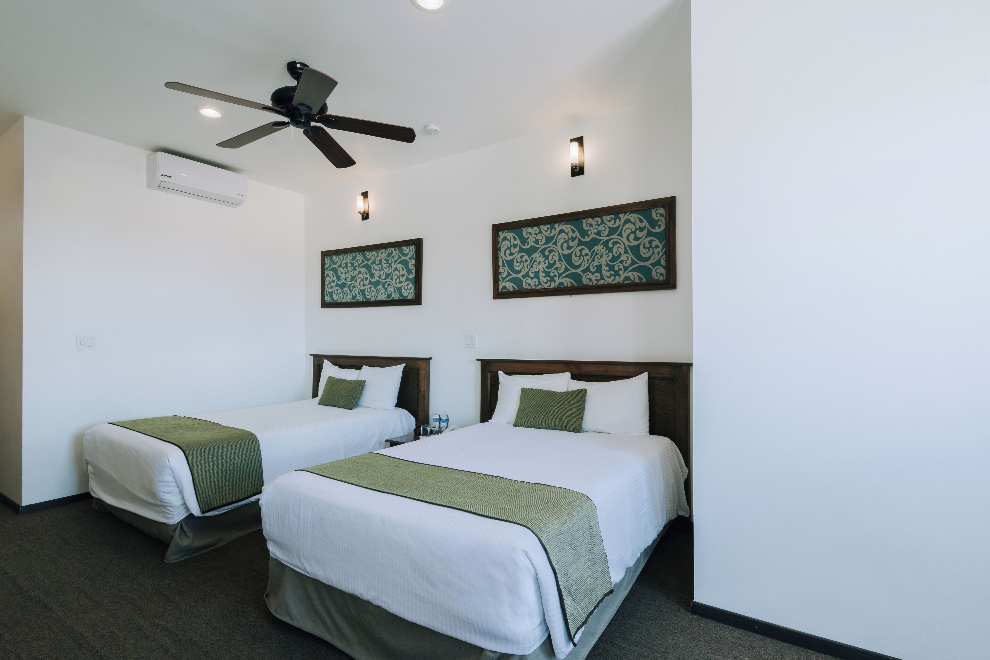 Une chambre d’hôtel avec deux lits, un ventilateur de plafond, un climatiseur mural et des œuvres d’art décoratives accrochées au mur.