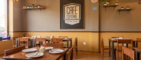 Interior de una cafetería con mesas y sillas de madera, un cartel enmarcado en la pared y lámparas colgantes.