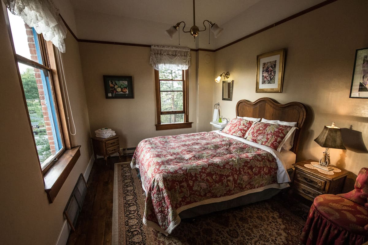 Un dormitorio con una cama grande, una mesita de noche, una silla, una ventana con cortinas de encaje y una lámpara de pared.