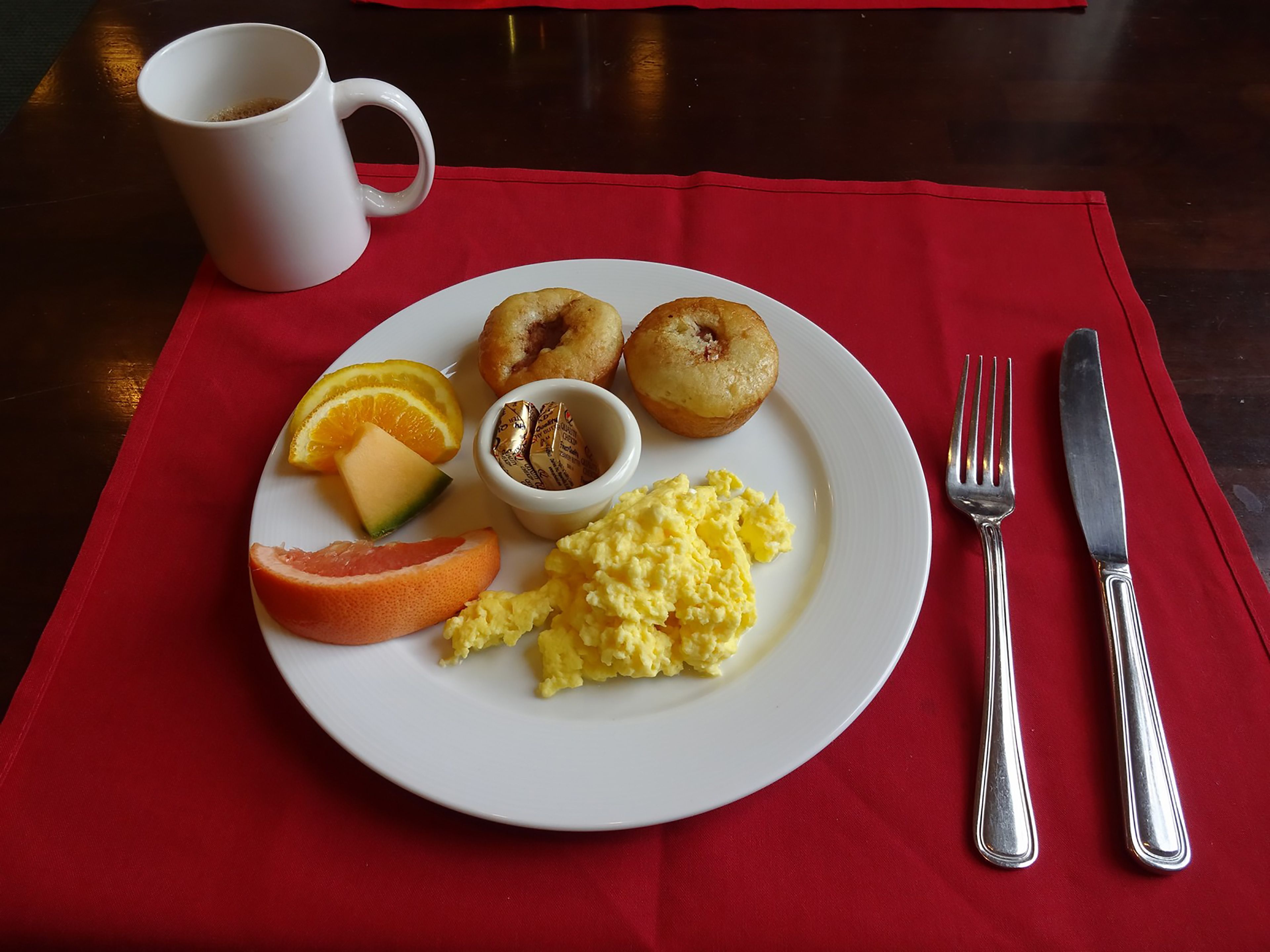 A breakfast plate with scrambled eggs, two pastries, orange slices, and a small container of butter on a red placemat.