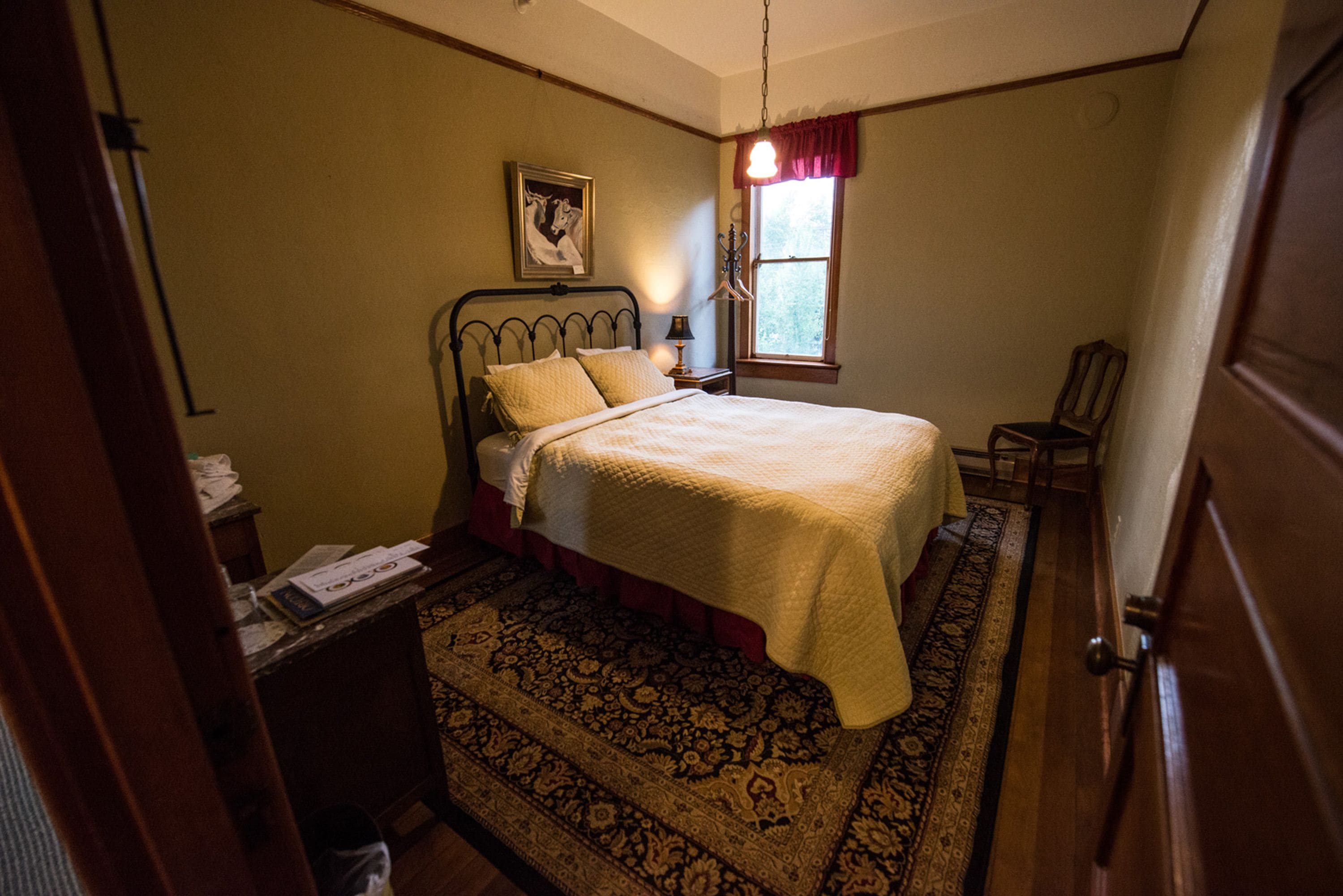 A bedroom with a bed, a nightstand, a chair, a window with a red valance, and a framed picture on the wall.