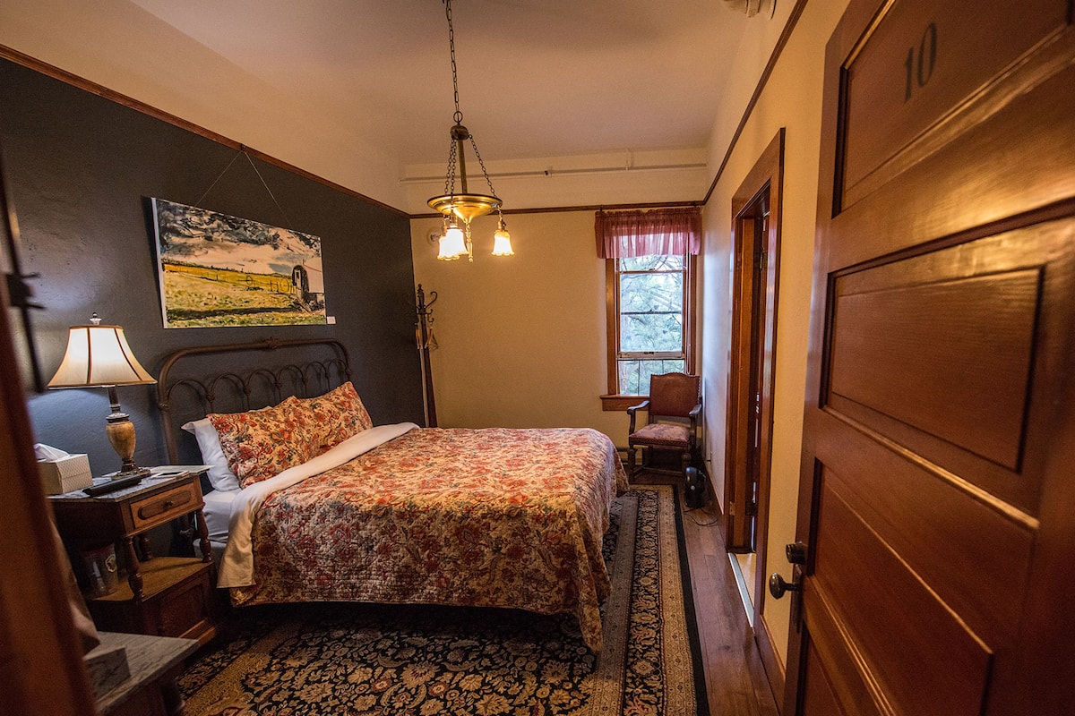 Un dormitorio con cama, mesita de noche, lámpara, silla, ventana y puerta.