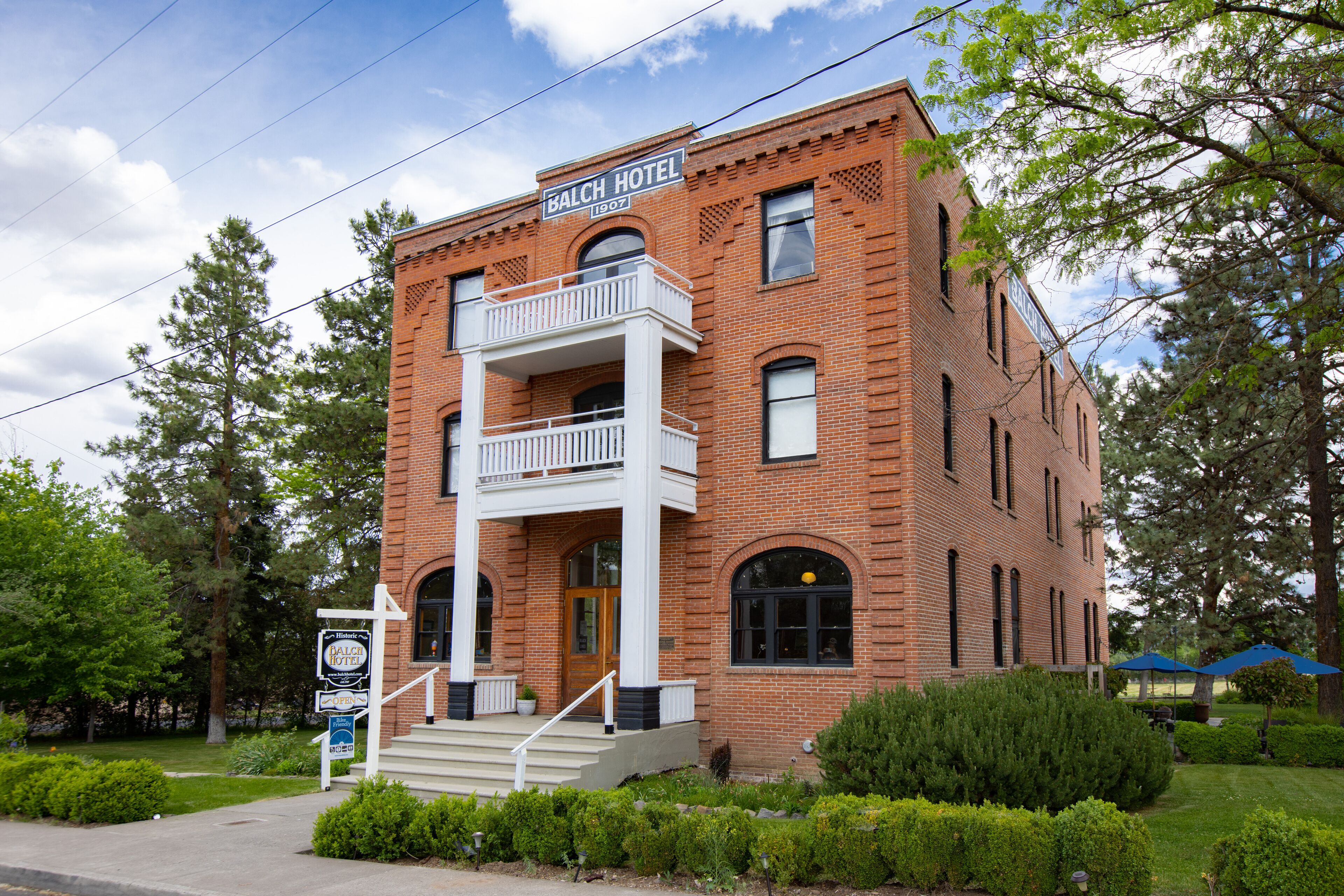A historic brick building with a balcony, labeled as the Balch Hotel from 1907, featuring a front entrance and steps leading up to it.