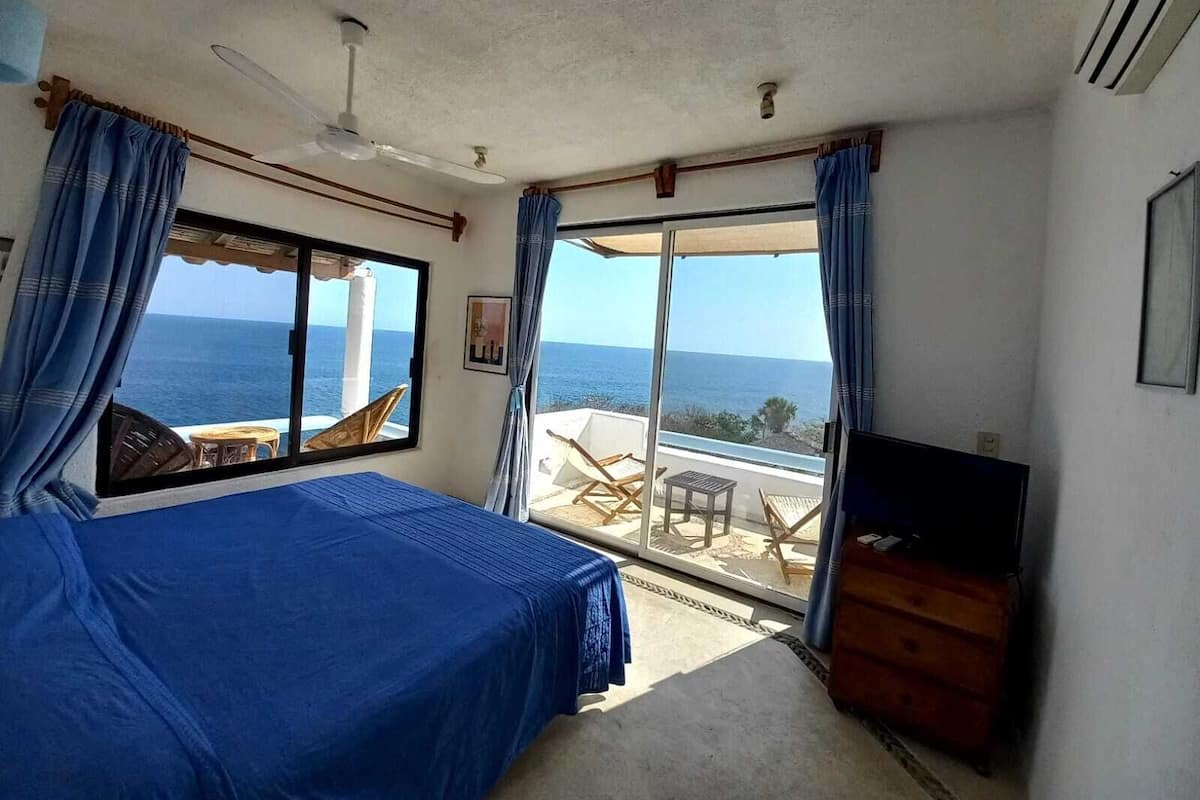 Een slaapkamer met een bed, een plafondventilator, een televisie en een balkon met uitzicht op zee.