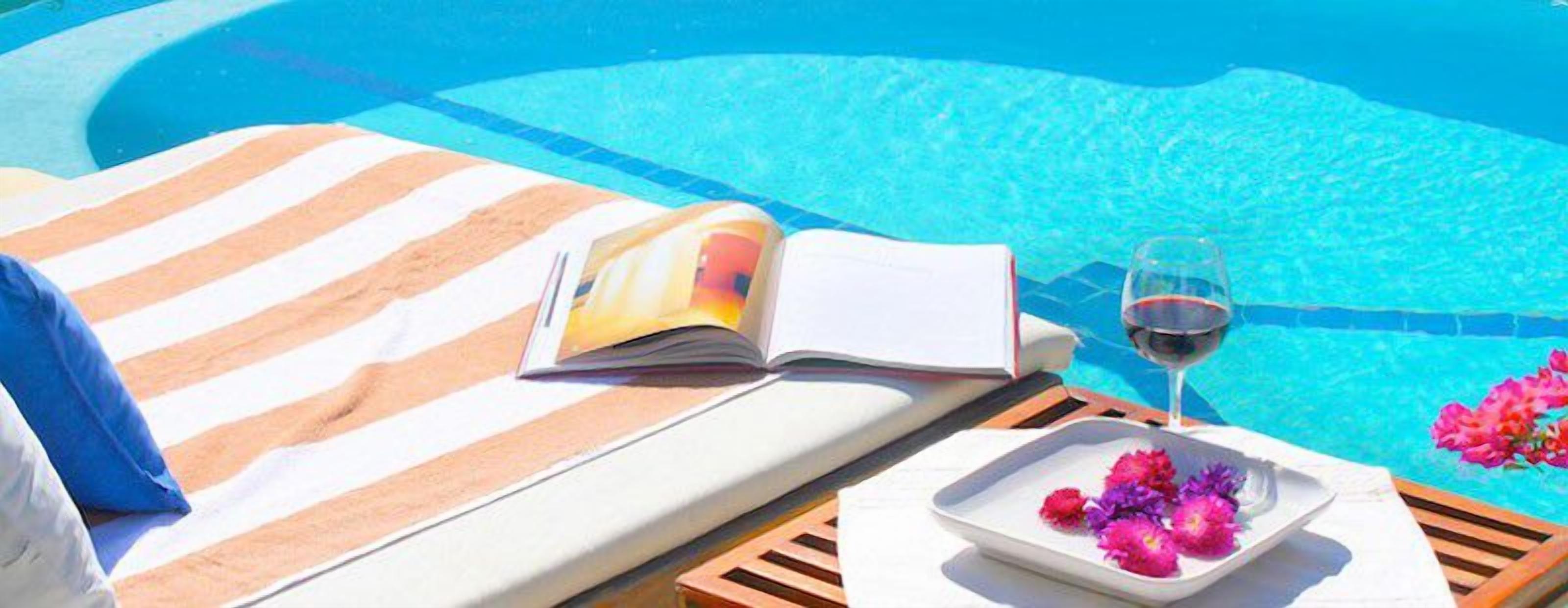 A poolside setting with a lounge chair, a book, a glass of red wine, and a plate of flowers.