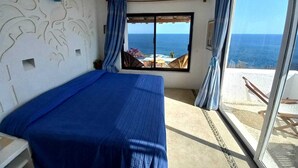 Una habitación con cama, ventilador de techo, ventana con cortinas y vista al océano.