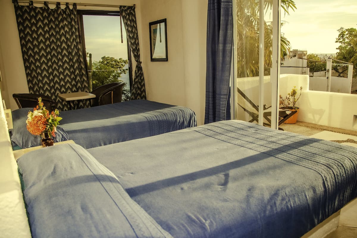 Een kamer met twee bedden, een klein tafeltje en een stoel. Er is een raam met gordijnen, een potplant en uitzicht op buiten.