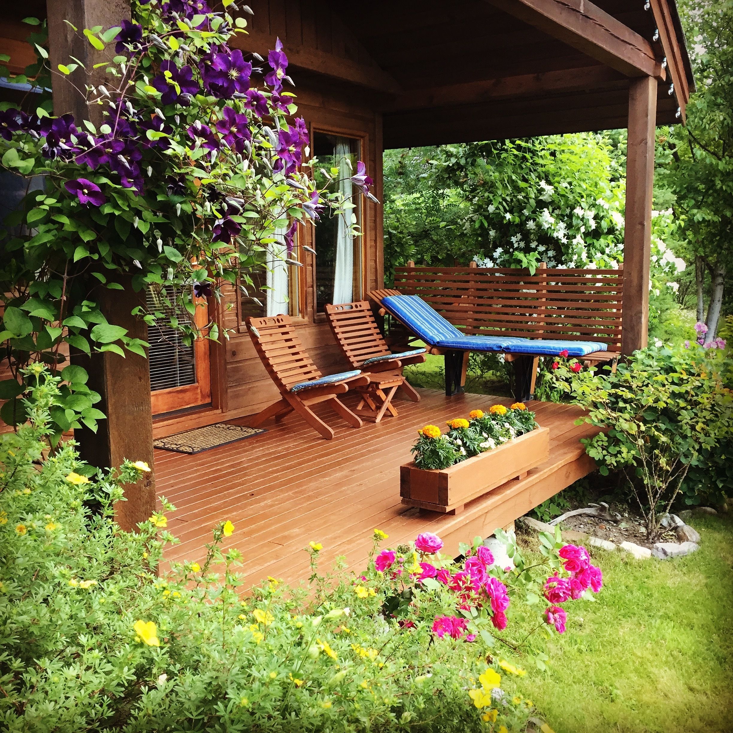 A wooden deck with two lounge chairs and a bench, surrounded by lush greenery and flowers.