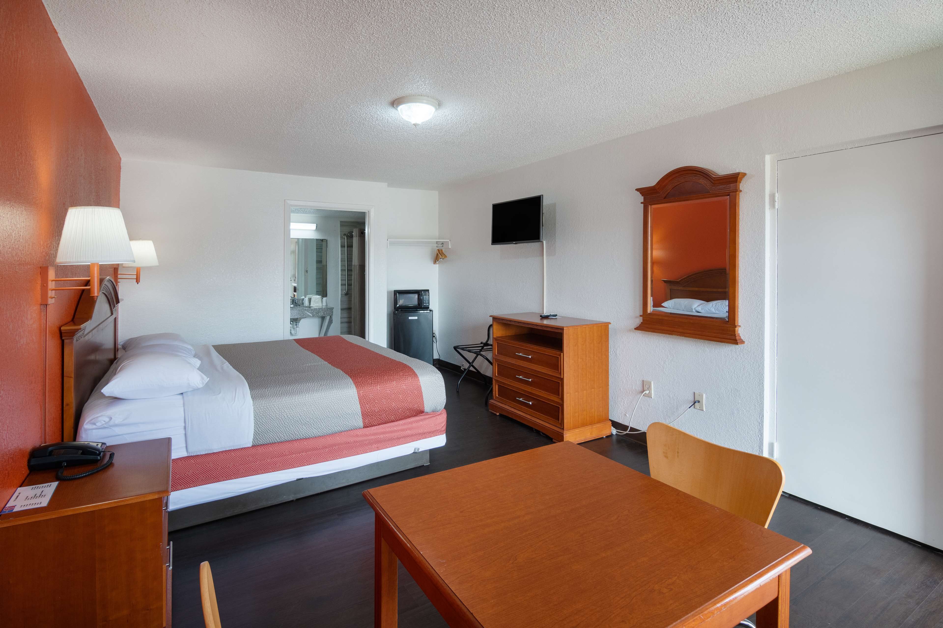 Quarto de hotel com cama, mesa de cabeceira, luminária, escrivaninha, cadeira, cômoda, televisão e porta.