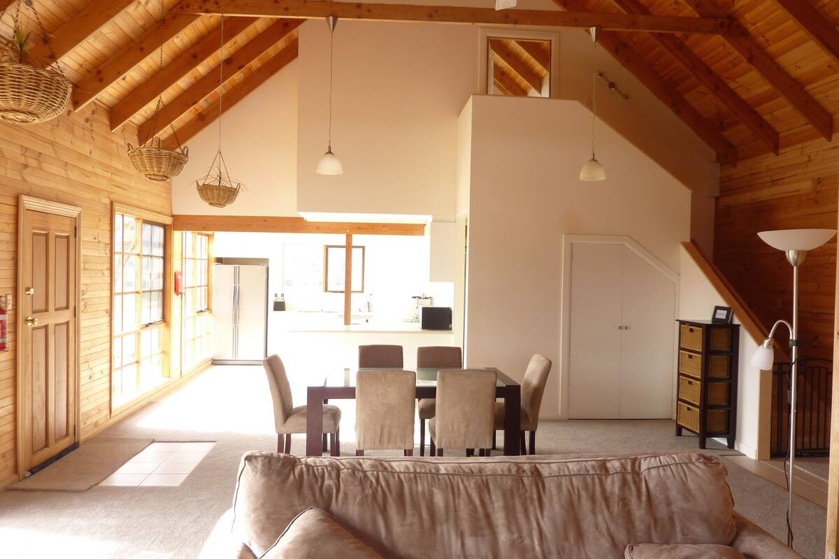 A spacious loft-style room with a high ceiling, wooden beams, a dining area with a table and chairs, and a cozy seating area with a sofa.