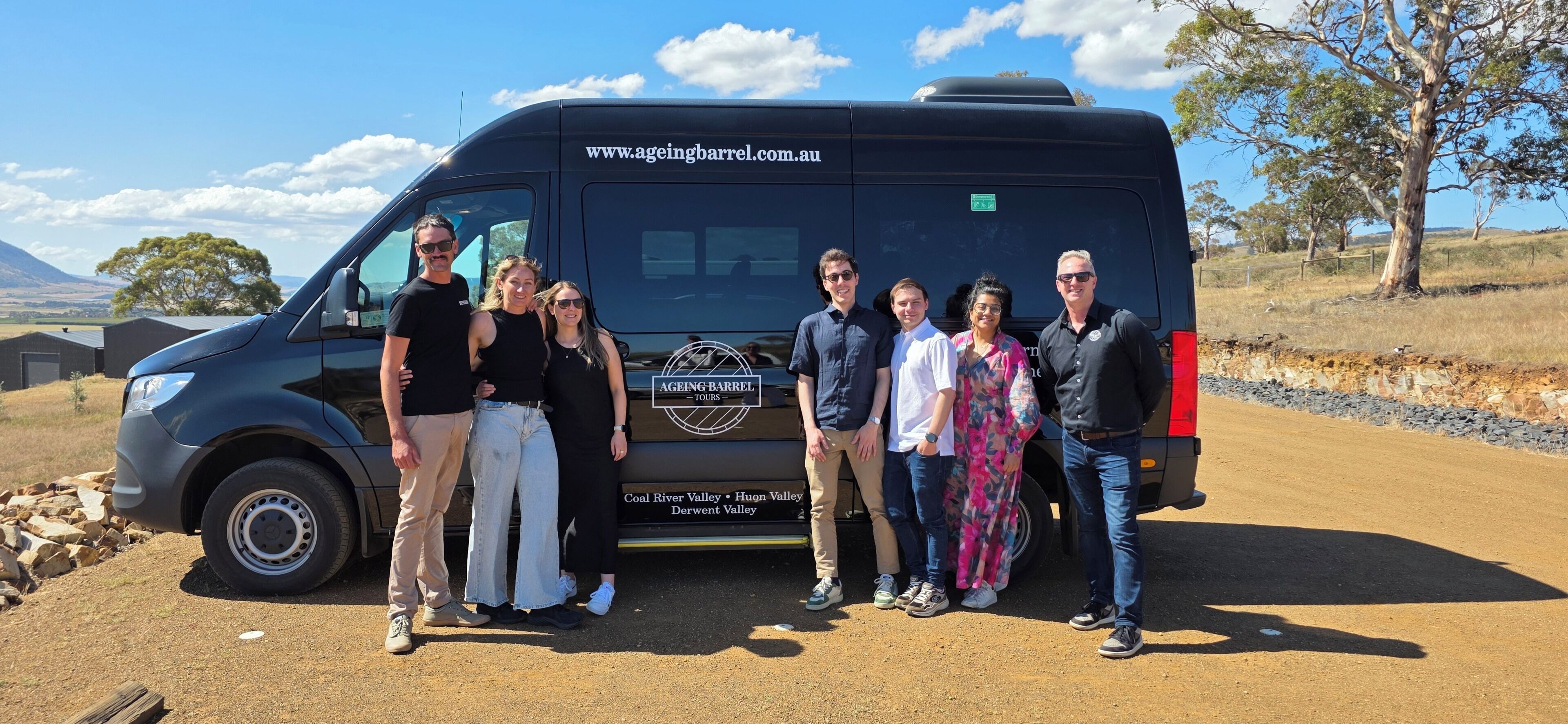 A group of people standing in front of a black van with a website address and tour locations on the back.