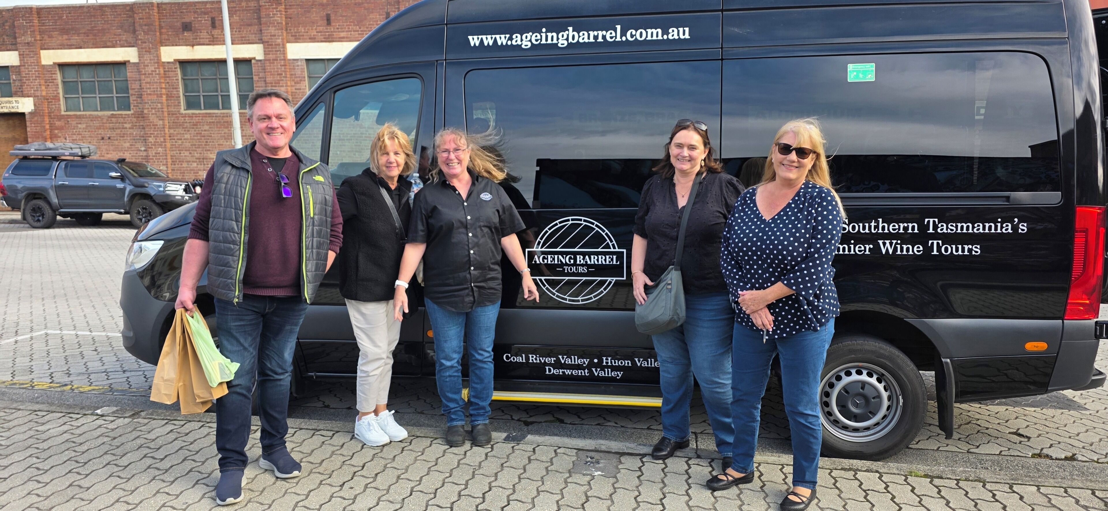A group of people standing in front of a black van with 'AGEING BARREL' branding and tour information.