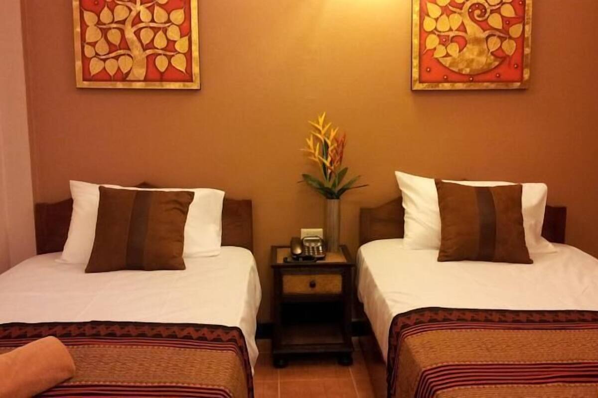 A hotel room with two beds, each with a brown pillow and a white pillow. There is a nightstand with a telephone and a vase with flowers between the beds.