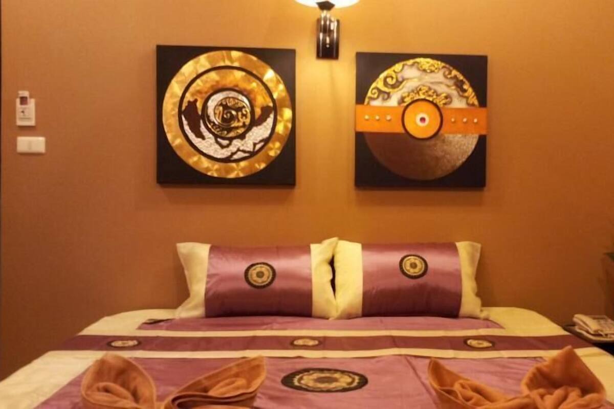 A hotel room with a bed, two wall-mounted artworks, a nightstand with a phone, and a potted plant.