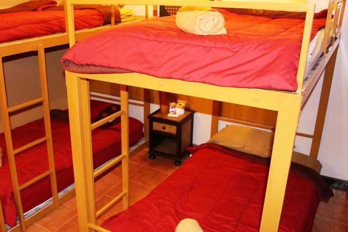 A dormitory room with bunk beds, each with red bedding. There is a small bedside table with a lamp and a bottle.