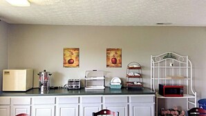 A kitchen with a refrigerator, toaster, coffee maker, microwave, and a white metal shelf. There are two paintings on the wall. - Sunrise Inn Williamstown (Williamstown)