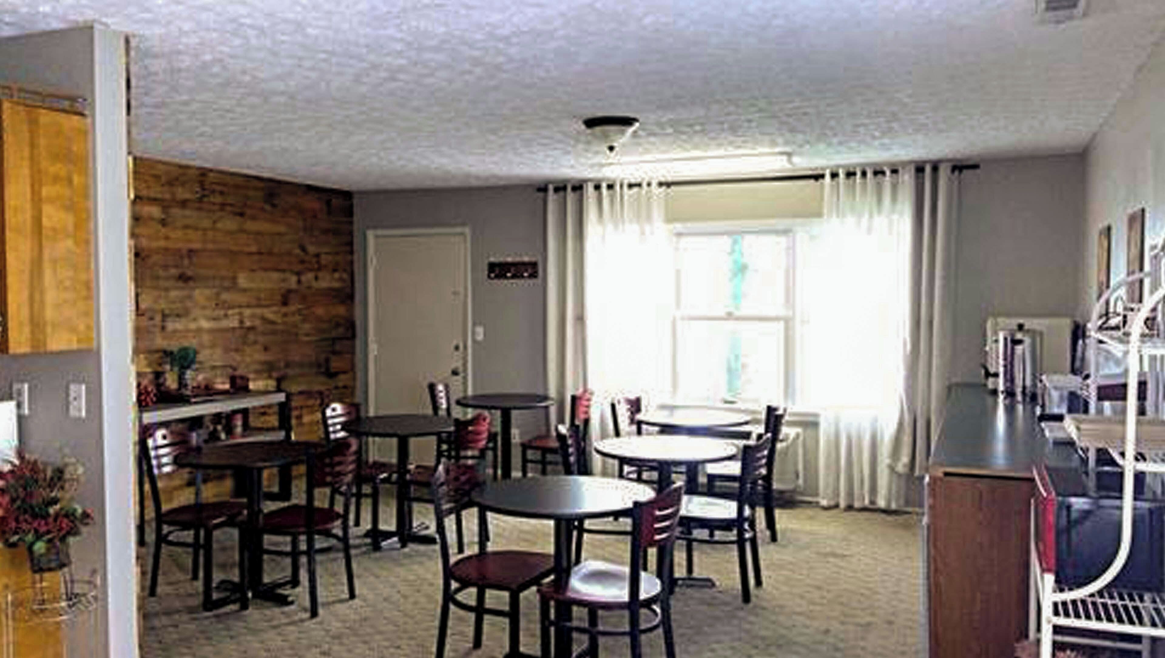 Comedor con varias mesas y sillas, pared de madera, una puerta y un ventanal con cortinas.