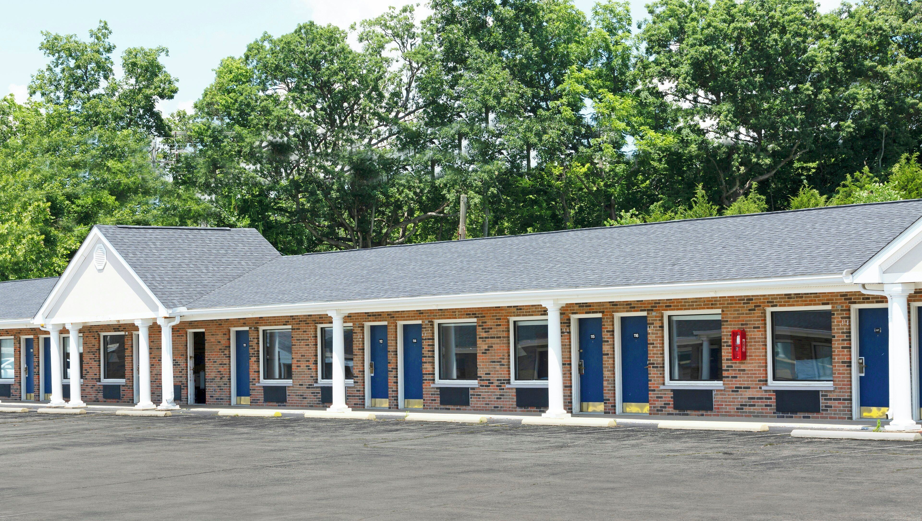 A row of brick motel rooms with blue doors, white columns, and a parking lot in front.