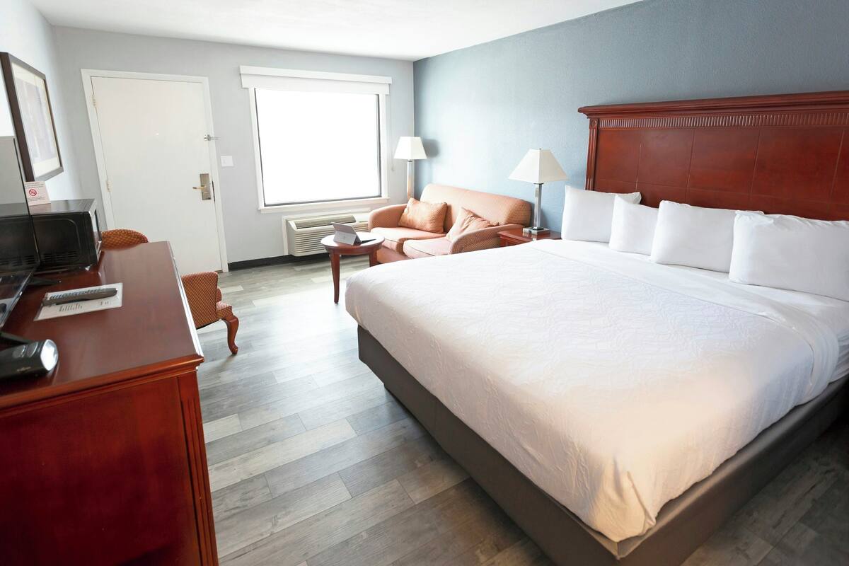 A hotel room with a bed, a desk with a computer, a chair, a lamp, and a window.
