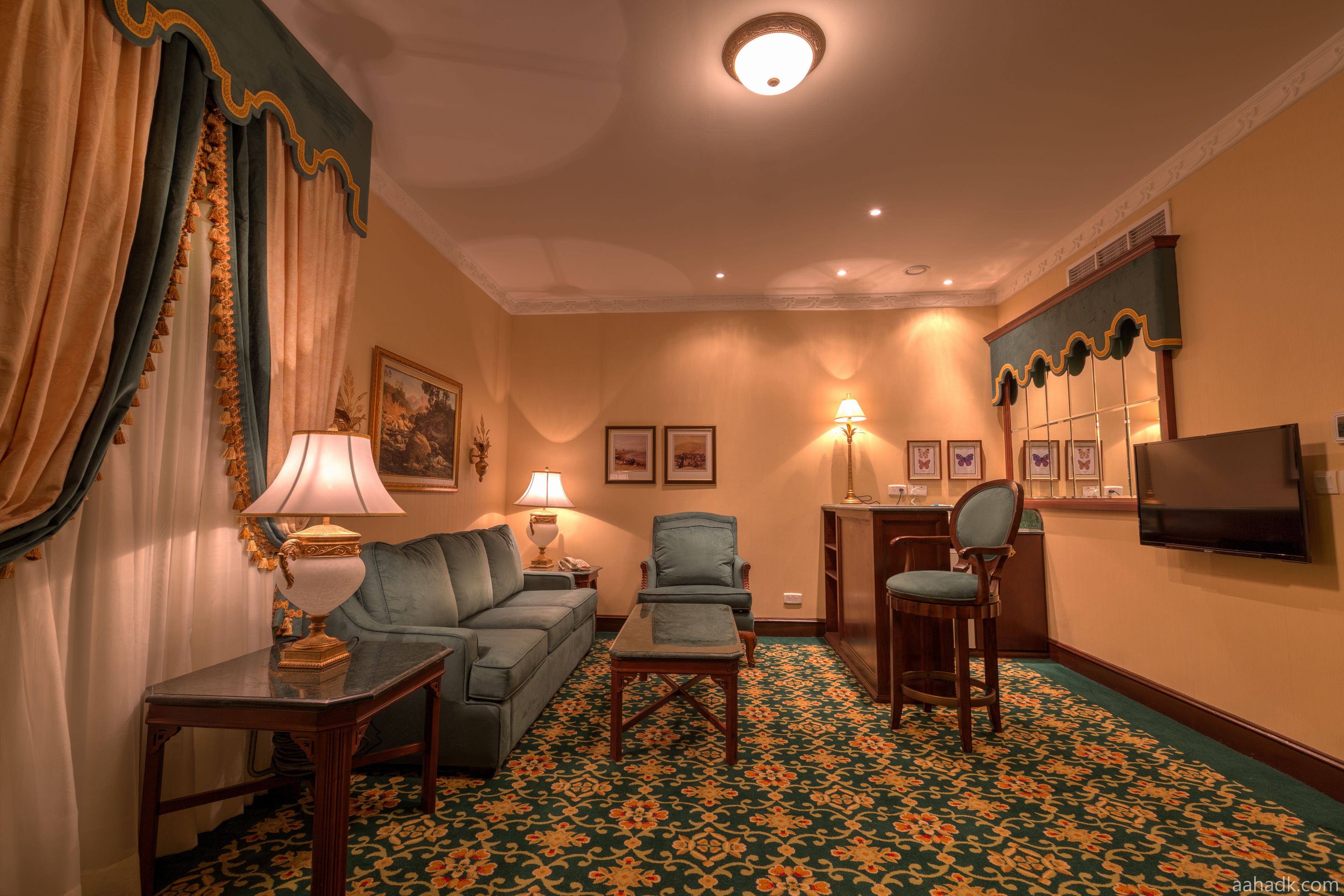 Une chambre d’hôtel avec un tapis à motifs verts, un canapé, un fauteuil, une petite table, une lampe et un téléviseur fixé au mur.
