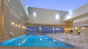 An indoor swimming pool with clear blue water, surrounded by lounge chairs and umbrellas, with large windows offering a view of the city at night.