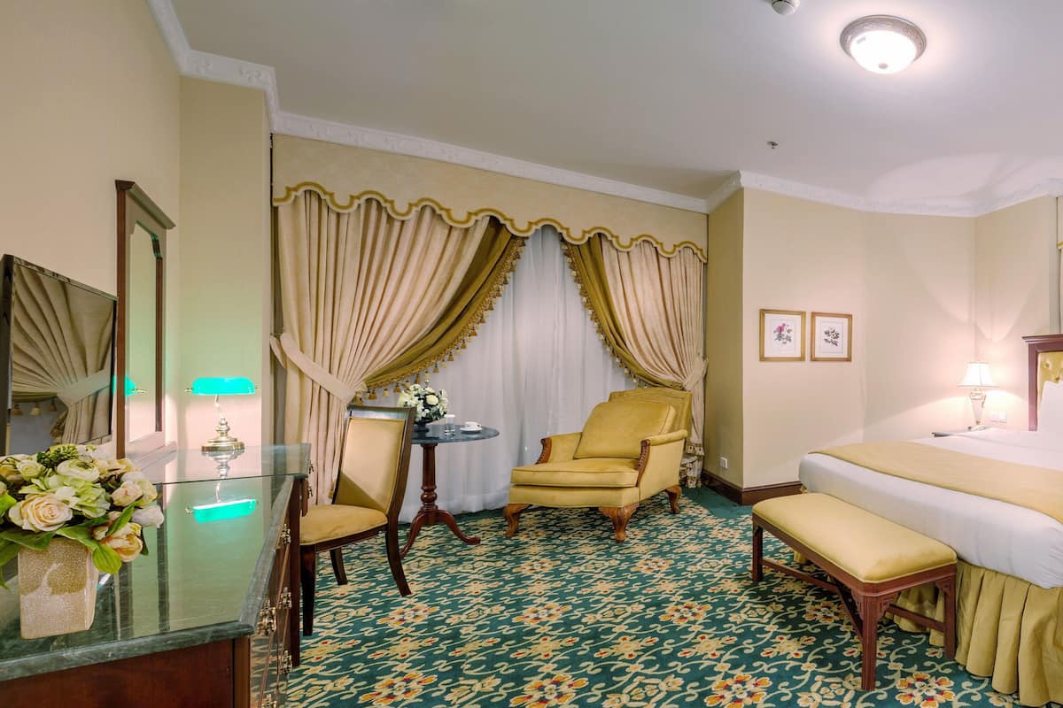 A hotel room with a large bed, a chair, a bench, a desk with a mirror, and a vase of flowers.