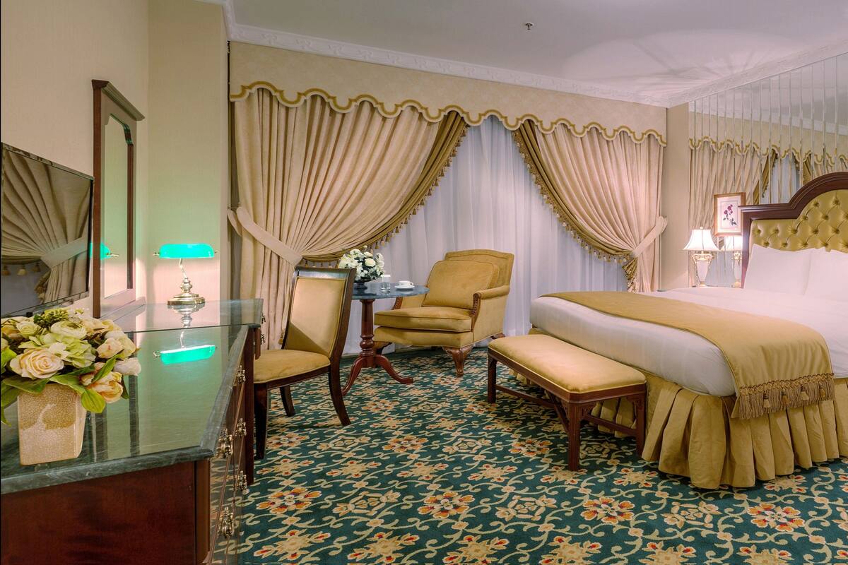 A hotel room with a large bed, a sitting area with a chair and a table, a desk with a lamp, and a mirror.