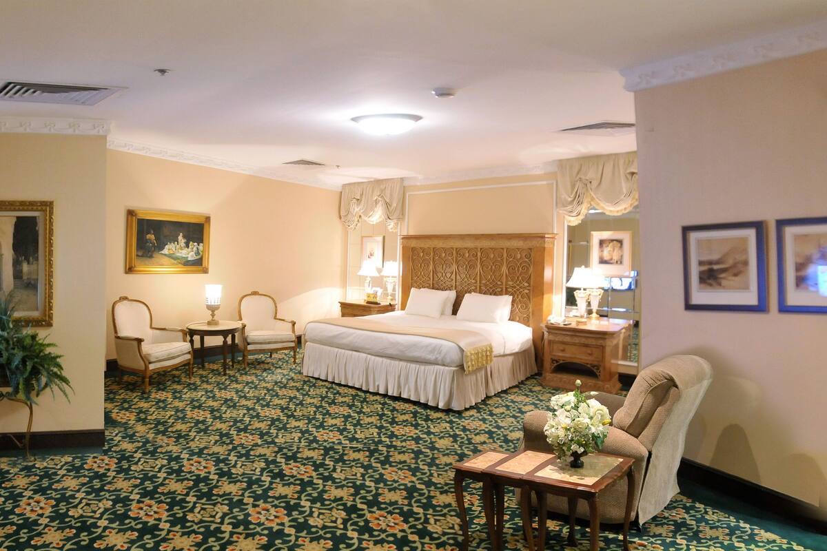 A hotel room with a large bed, a sitting area with a chair and table, a desk with a computer, and a large mirror.