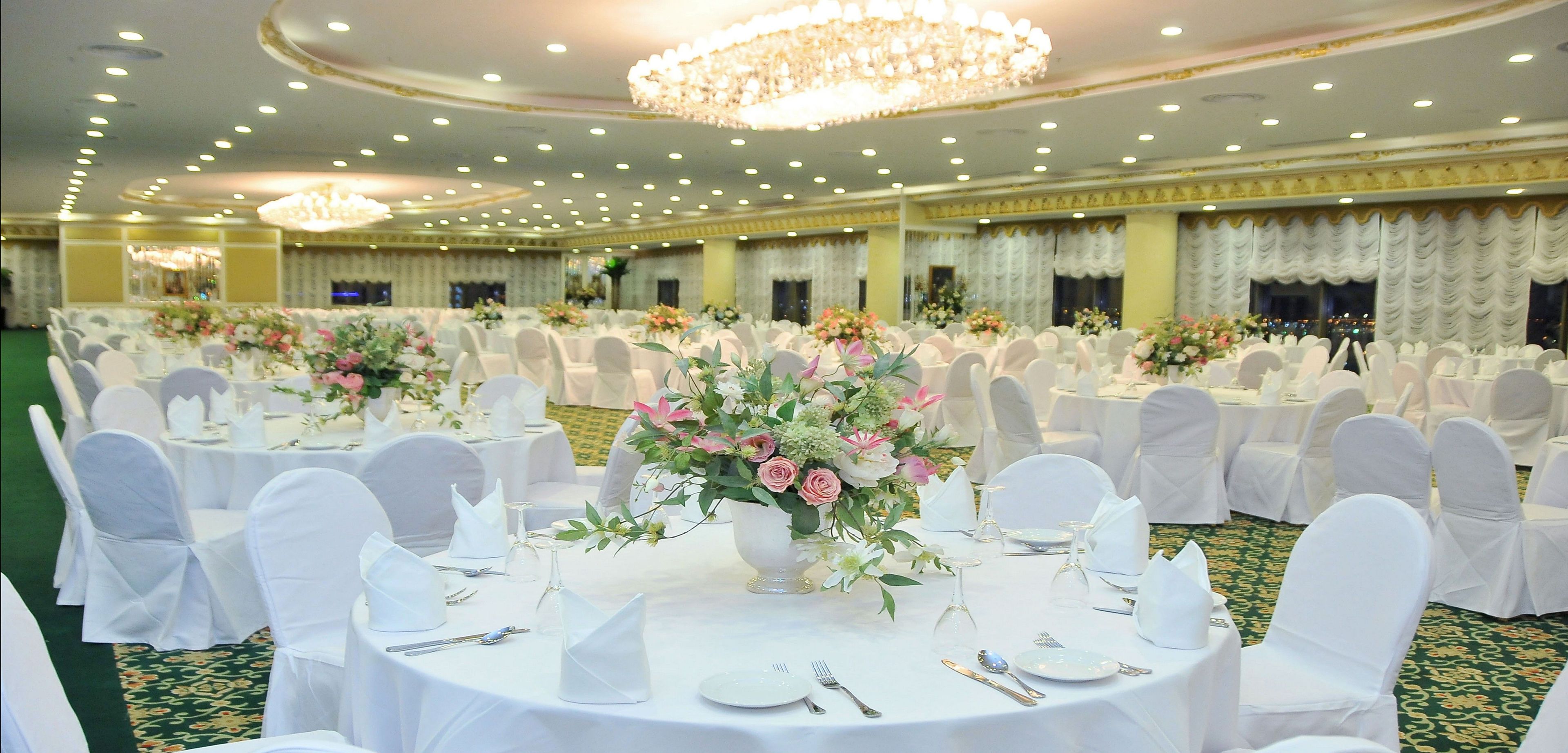 Un salón de banquetes con mesas redondas, sillas blancas y centros de mesa florales.