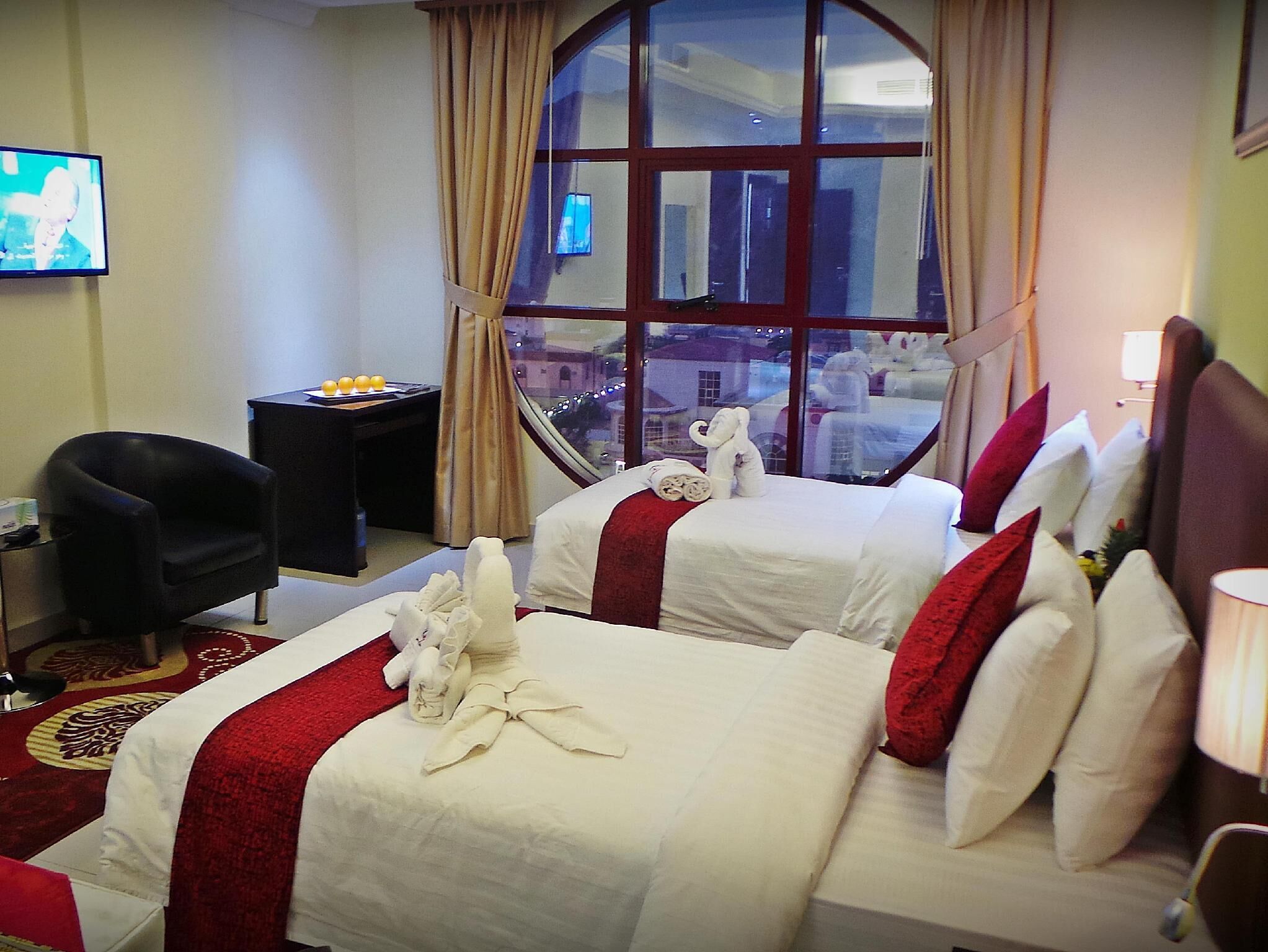 A hotel room with two beds, a TV, a desk with a lamp, and a view of the city through a large window.
