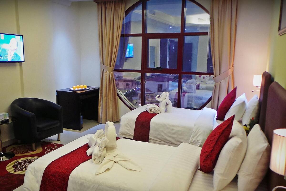 A hotel room with two beds, a desk, a chair, a TV, and a large window with curtains.