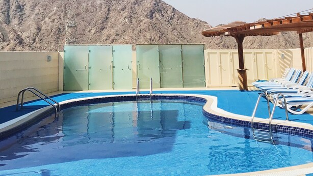 A swimming pool with a view of rocky mountains, a covered area with lounge chairs, and a metal ladder. - Mirage Hotel Al Aqah (Dhadnah)