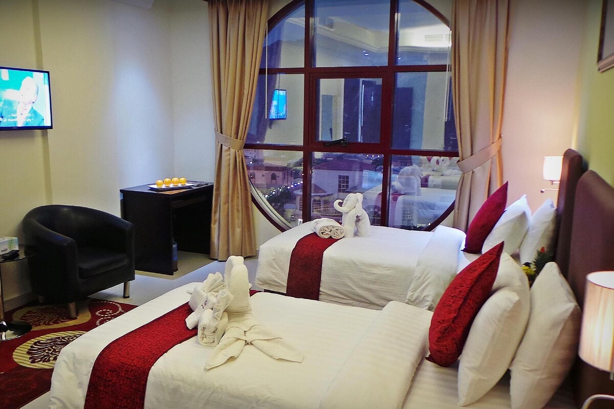A hotel room with two beds, a TV, a desk, and a window with a city view.
