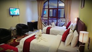 A hotel room with two beds, a TV, a chair, a small table with a tray of fruit, and a window with a view of buildings. - Mirage Hotel Al Aqah (Dhadnah)