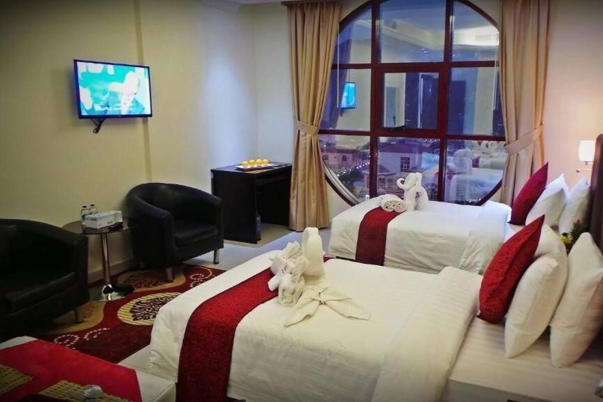 A hotel room with two beds, a TV, a chair, a small table with a tray of fruit, and a window with a view of buildings.
