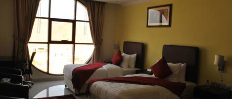 Habitación de hotel con dos camas, un ventanal con cortinas, un cuadro en la pared y un escritorio con lámpara.