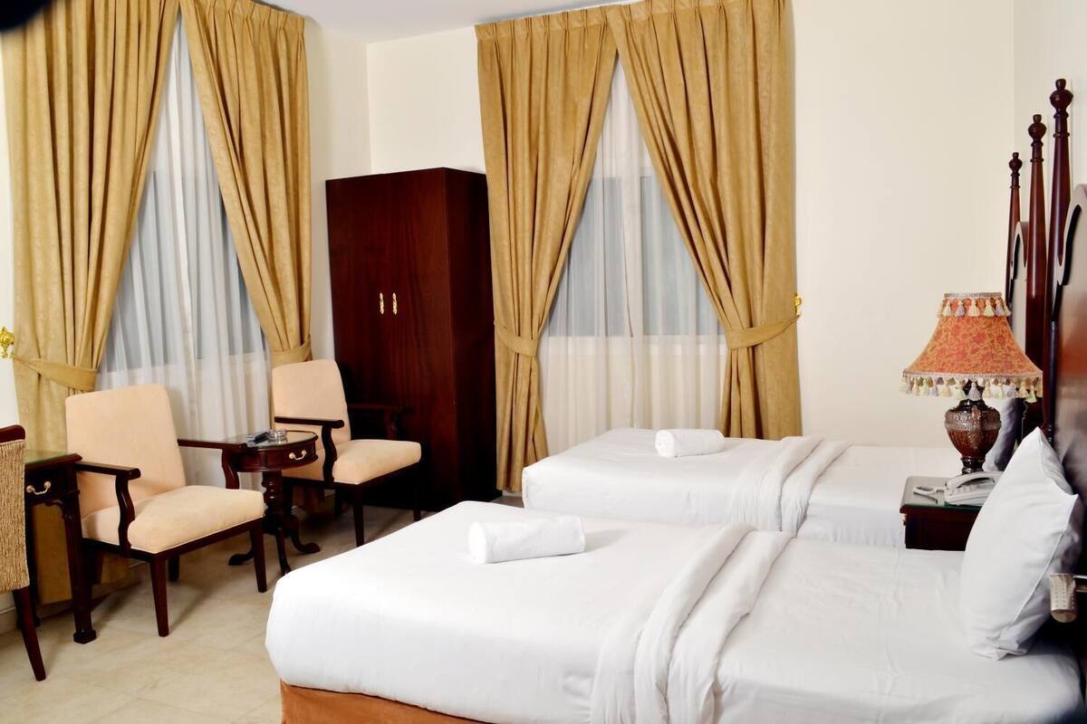 A hotel room with two beds, a wooden wardrobe, a desk with a chair, and a lamp.